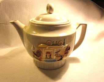 May include: A white ceramic teapot with a brown and gold design of a building with a chimney and a window. The teapot has a lid with a knob on top.
