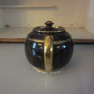 May include: A brown teapot with a gold handle and trim. The teapot has a round body and a lid with a knob on top.