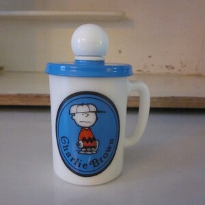 Puede incluir: Taza de cerámica blanca con tapa azul y una imagen de dibujos animados de Charlie Brown con una gorra de béisbol y sosteniendo un bate de béisbol. La taza tiene el texto "Charlie Brown" alrededor de la imagen.