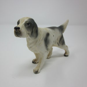 May include: A white and gray porcelain figurine of a dog, possibly a pointer breed, standing with its head tilted to the side. The dog has black eyes and a black nose.