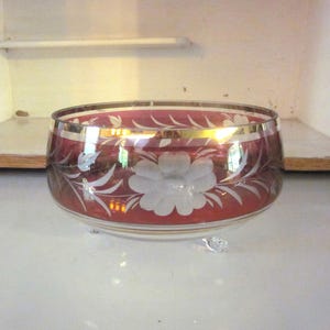 Puede incluir: Un cuenco de vidrio rojo con un borde dorado y un diseño floral blanco. El cuenco tiene tres patas y es perfecto para servir aperitivos o fruta.