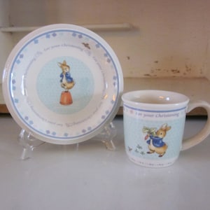 Puede incluir: Un juego de taza y platillo de cerámica azul y blanco con un diseño de Peter Rabbit. La taza tiene un asa y el platillo tiene un borde festoneado. El texto "For your Christening" está impreso en la taza.