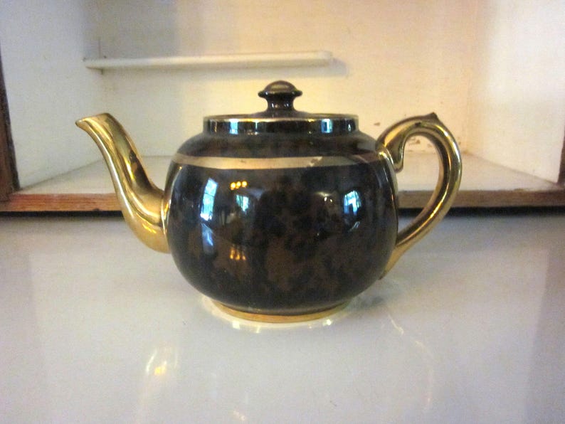 May include: A black teapot with gold trim and a speckled design. The teapot has a gold spout and handle.