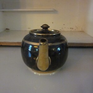 May include: A black teapot with a gold band around the top and a gold spout. The teapot is sitting on a white surface.