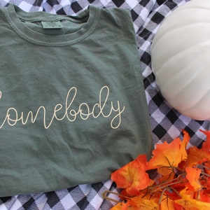 Puede incluir: Una camiseta verde con la palabra "homebody" escrita en blanco en la parte delantera. La camiseta est&aacute; sobre una tela a cuadros blanco y negro con una calabaza blanca y hojas de oto&ntilde;o naranjas.