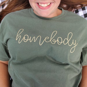 Puede incluir: Una camiseta verde con la palabra "homebody" escrita en letras doradas.