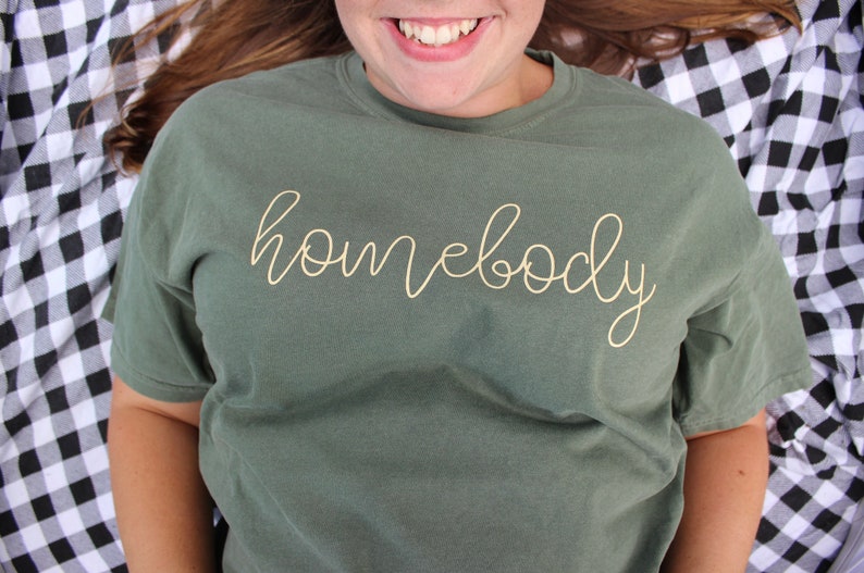 Puede incluir: Una camiseta verde con la palabra "homebody" escrita en letras doradas.