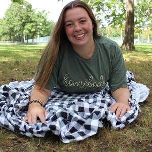 Puede incluir: Una joven con una camiseta verde con el texto "homebody" en letras amarillas, sentada en una manta a cuadros negros y blancos en un parque de c&eacute;sped.