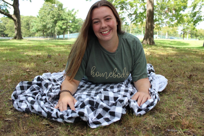 Puede incluir: Una joven con una camiseta verde con el texto "homebody" en letras amarillas, sentada en una manta a cuadros negros y blancos en un parque de c&eacute;sped.