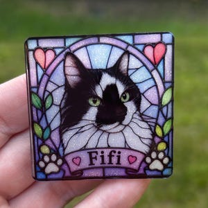 May include: A square decorative plaque featuring a black and white cat portrait centered within a stained glass style frame. The design includes purple and blue tones, pink hearts, green leaves, white paw prints, and the name Fifi in black text.