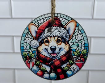 May include: A round Christmas ornament featuring a cartoon Corgi dog wearing a Santa hat and a red and black plaid scarf. The ornament has a stained glass design with colorful ornaments and holly.