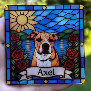 May include: A square coaster featuring a stained glass style illustration of a tan and white dog. The design includes a yellow sun, blue water, green hills, and red roses on both sides. A white banner at the bottom displays the name Axel in black.
