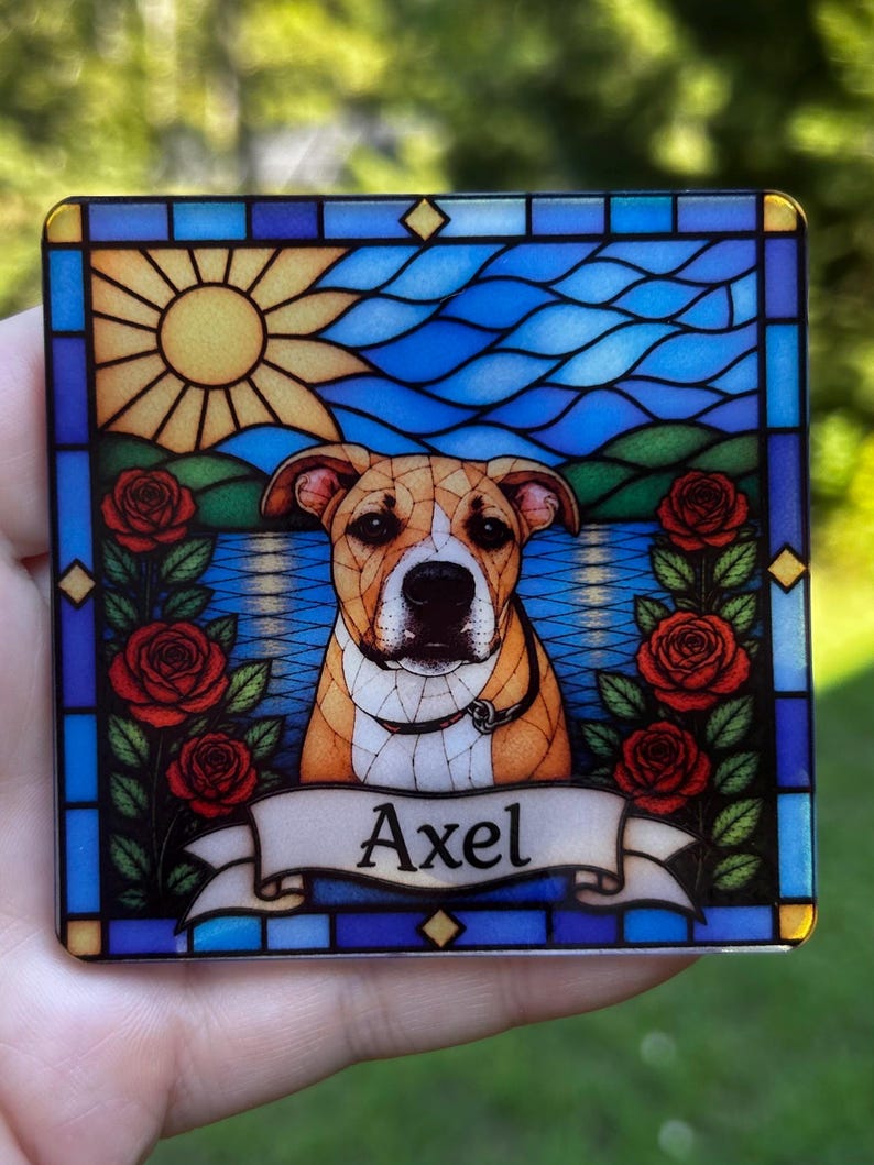 May include: A square coaster featuring a stained glass style illustration of a tan and white dog. The design includes a yellow sun, blue water, green hills, and red roses on both sides. A white banner at the bottom displays the name Axel in black.