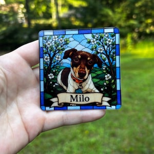 May include: A square coaster featuring a stained glass style illustration of a brown and white dog with an orange collar. The dog is framed by flowering trees and a blue border. A cream-coloured banner at the bottom displays the name Milo in black.