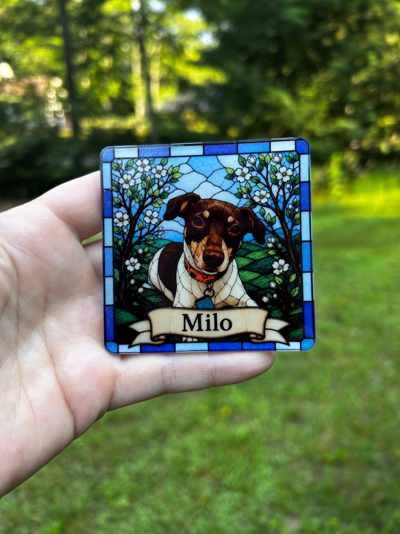 May include: A square coaster featuring a stained glass style illustration of a brown and white dog with an orange collar. The dog is framed by flowering trees and a blue border. A cream-coloured banner at the bottom displays the name Milo in black.