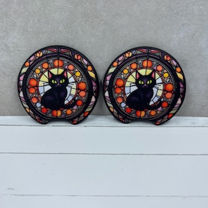 May include: Two round car coasters featuring a black cat design within a stained-glass style frame. The coasters have a black cat silhouette with yellow eyes, surrounded by orange, yellow, and red colors. The background is a light gray.