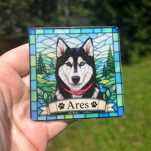 May include: A square coaster featuring a stained glass style illustration of a black and white husky with heterochromia, wearing a red collar. The background shows a landscape with trees and a river. A banner at the bottom displays the name Ares.