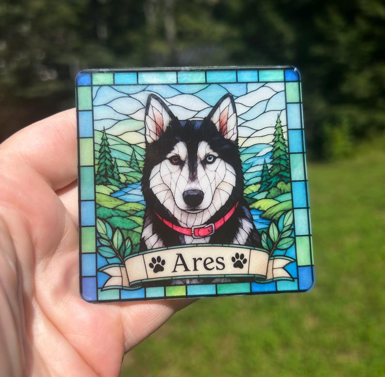 May include: A square coaster featuring a stained glass style illustration of a black and white husky with heterochromia, wearing a red collar. The background shows a landscape with trees and a river. A banner at the bottom displays the name Ares.
