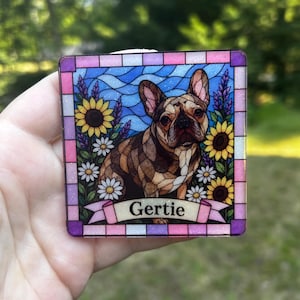 May include: A square decorative plaque featuring a stained glass style illustration of a brown French Bulldog surrounded by sunflowers and daisies against a blue wavy background. A pink banner at the bottom displays the name Gertie in black text.