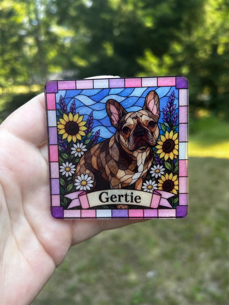 May include: A square decorative plaque featuring a stained glass style illustration of a brown French Bulldog surrounded by sunflowers and daisies against a blue wavy background. A pink banner at the bottom displays the name Gertie in black text.