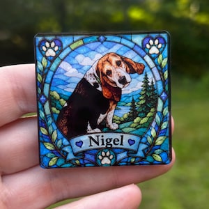 May include: A square magnet featuring a stained glass style illustration of a brown, black, and white beagle dog. The dog is centred within a blue circular frame surrounded by green foliage, with the name Nigel printed on a banner at the bottom.