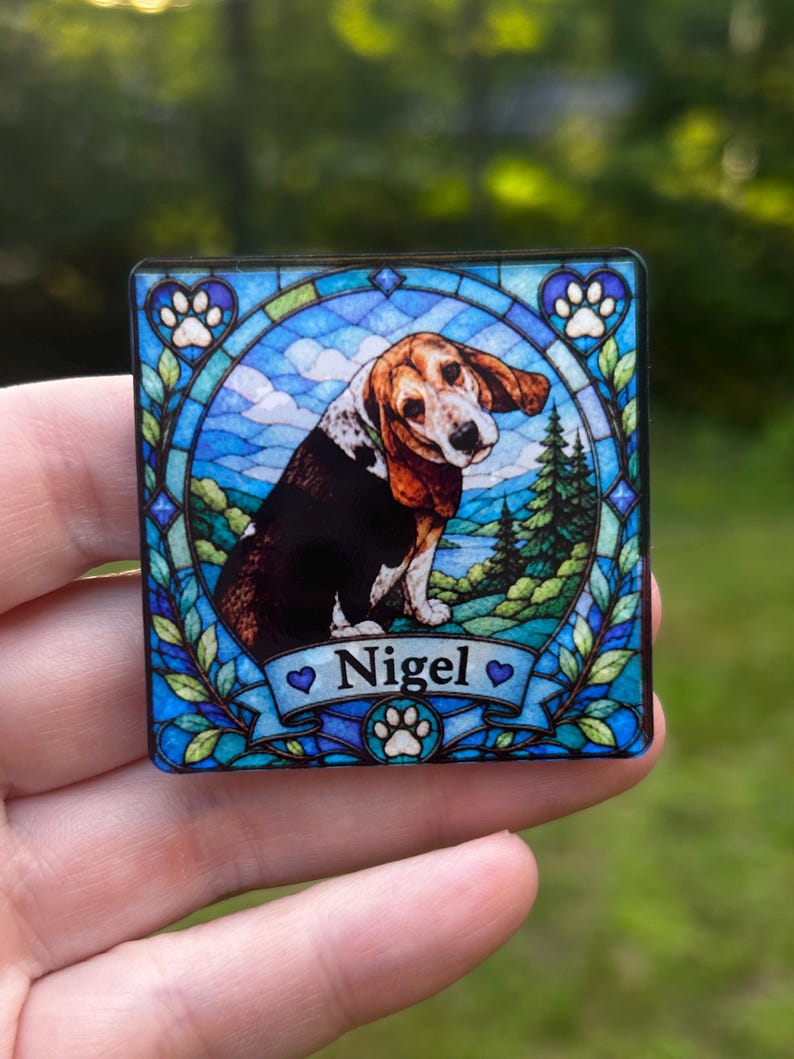 May include: A square magnet featuring a stained glass style illustration of a brown, black, and white beagle dog. The dog is centred within a blue circular frame surrounded by green foliage, with the name Nigel printed on a banner at the bottom.