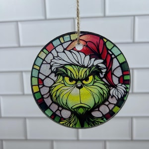 May include: A round Christmas ornament featuring a green character with an angry expression wearing a red and white Santa hat. The ornament has a stained glass effect with a colorful border and is suspended by a twine string.