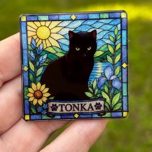 May include: A square decorative plaque featuring a stained glass style illustration of a black cat sitting in a garden with a yellow sun, blue flowers, and green foliage. A banner at the bottom displays the name TONKA with paw print accents.