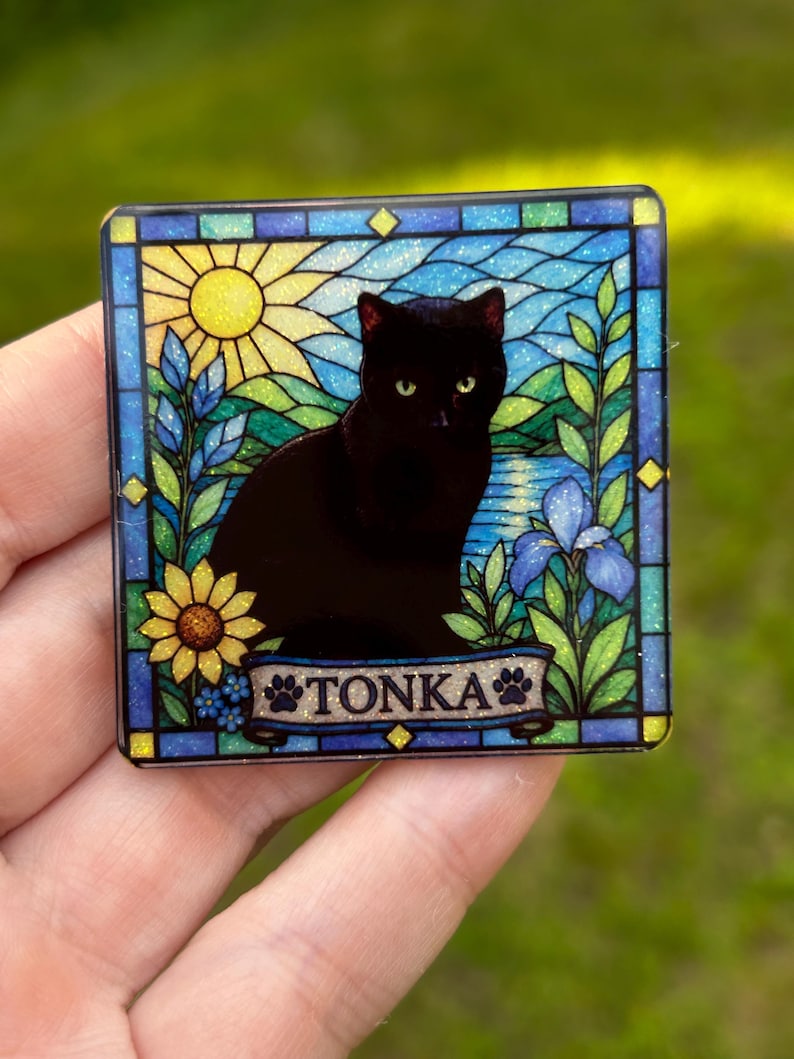 May include: A square decorative plaque featuring a stained glass style illustration of a black cat sitting in a garden with a yellow sun, blue flowers, and green foliage. A banner at the bottom displays the name TONKA with paw print accents.