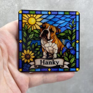 May include: A square coaster featuring a stained glass style illustration of a brown and white bulldog. The scene includes a yellow sun, blue water, green foliage, and a sunflower. A banner at the bottom displays the name Hanky in black text.