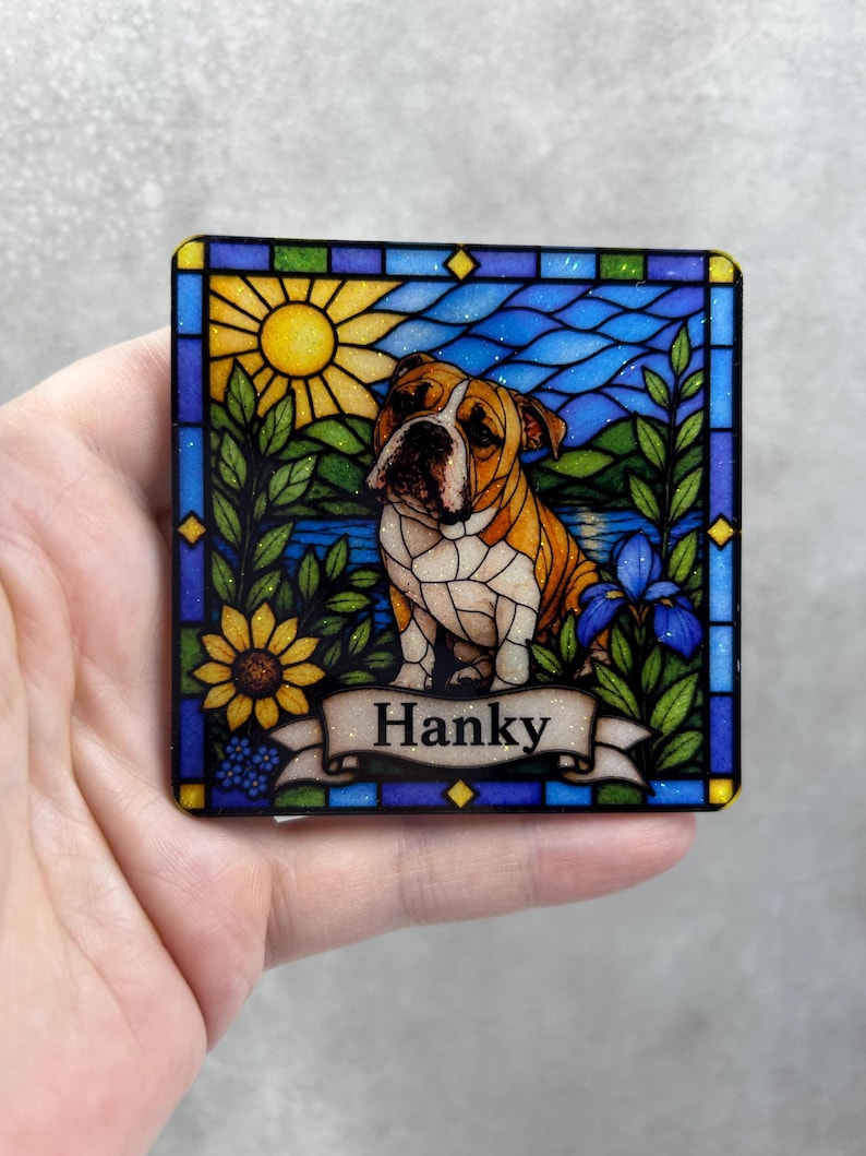 May include: A square coaster featuring a stained glass style illustration of a brown and white bulldog. The scene includes a yellow sun, blue water, green foliage, and a sunflower. A banner at the bottom displays the name Hanky in black text.