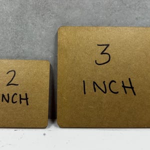 May include: Two square brown cardboard pieces with rounded corners. The smaller piece has the text 2 INCH written in black marker, and the larger piece has the text 3 INCH written in black marker, indicating their respective sizes in inches.