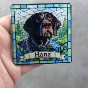 May include: A square coaster featuring a stained glass style illustration of a brown and white dog wearing a collar. The background depicts a landscape with green trees and a blue river. A banner at the bottom contains the name Hanz in black text.