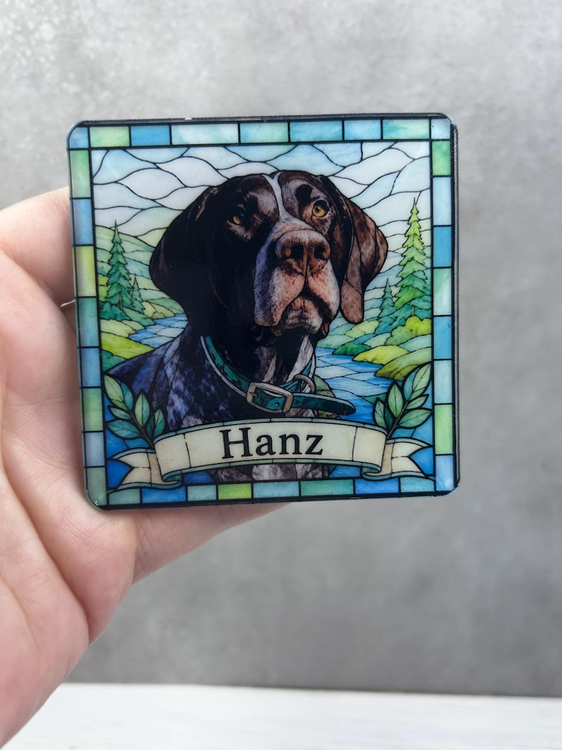 May include: A square coaster featuring a stained glass style illustration of a brown and white dog wearing a collar. The background depicts a landscape with green trees and a blue river. A banner at the bottom contains the name Hanz in black text.