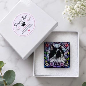 May include: A square pin featuring a black and white cat illustration in a stained glass style frame with the name Fifi printed on a banner. The pin is inside a white gift box with a Simply Ryn sticker that reads A small keepsake. A lifetime of memories.