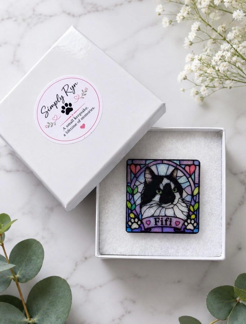 May include: A square pin featuring a black and white cat illustration in a stained glass style frame with the name Fifi printed on a banner. The pin is inside a white gift box with a Simply Ryn sticker that reads A small keepsake. A lifetime of memories.