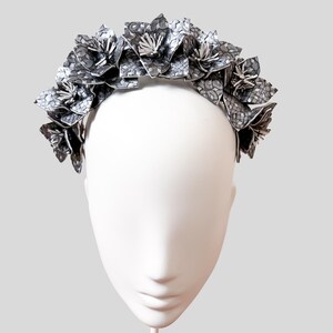 Può includere: Una corona floreale con fiori in tessuto a fantasia grigia e bianca. I fiori sono disposti attorno a una fascia, creando un copricapo decorativo. La corona è esposta su una testa di manichino bianca.