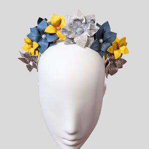 Pode incluir: Uma coroa de flores decorativa com flores de couro prateadas, amarelas e azuis. As flores são dispostas em torno de uma faixa de cabeça prateada. Pequenos detalhes de pérolas são colocados entre as flores. A coroa é exibida em uma cabeça de manequim branca.