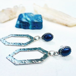 May include: A pair of silver earrings with a geometric hexagon shape and a dark blue gemstone dangle. The earrings have a hammered texture.