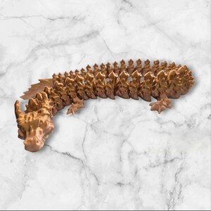 Könnte beinhalten: Eine goldfarbene, 3D-gedruckte Drachenfigur mit einem langen, segmentierten Körper und einem detaillierten Kopf. Der Drache ist zu einer Spirale zusammengerollt, mit dem Kopf auf dem Schwanz.