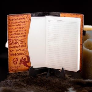 May include: Open leather journal with lined pages, displayed on a black stand. The cover is brown with dark red embossed text and symbols. The journal is open to reveal blank pages with a "Page" and "Content" section.