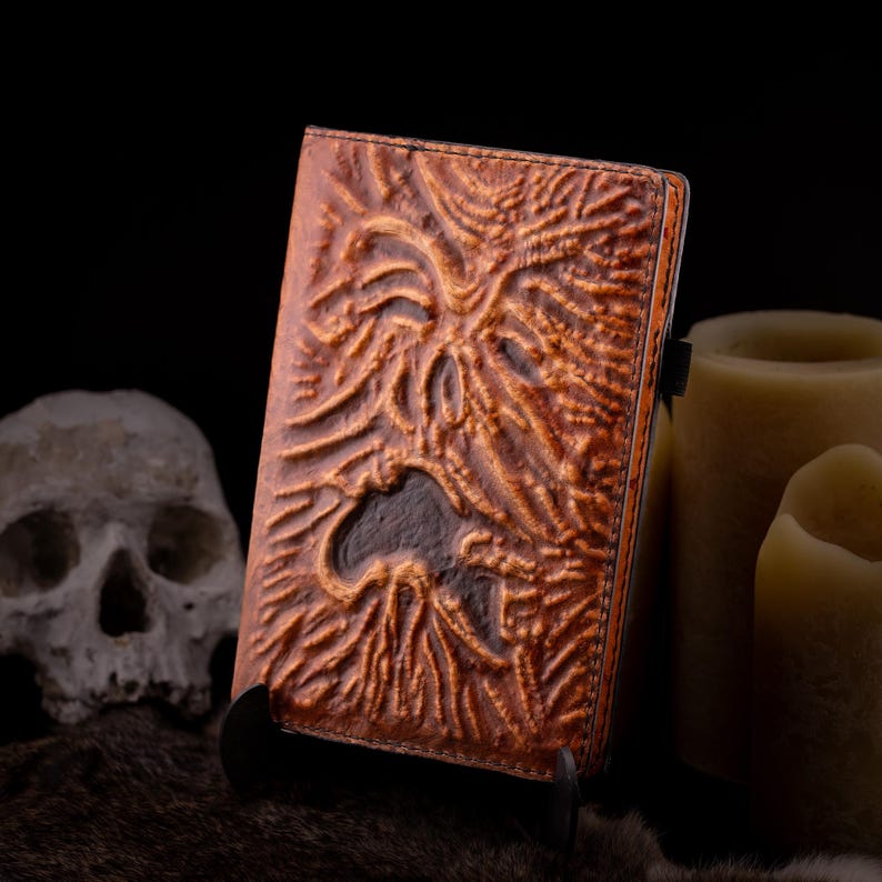 May include: A brown leather-bound book with a detailed, embossed design featuring a grotesque face. The book is propped up on a black stand, with a skull and candles in the background, creating a spooky, gothic aesthetic.
