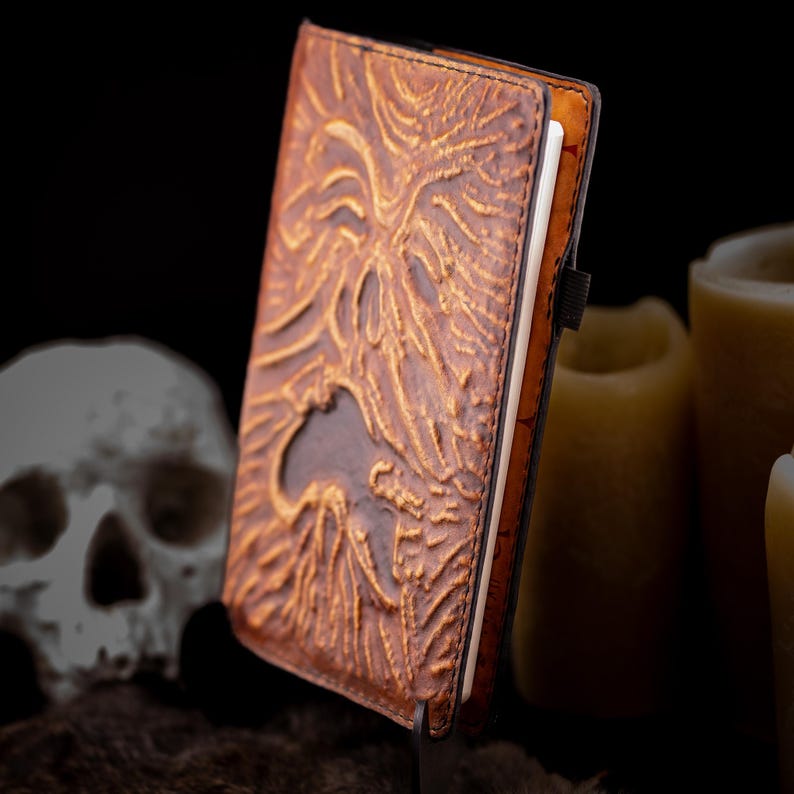 May include: A brown leather journal with an embossed design. The cover features a detailed, textured pattern with a central cutout. The journal is closed with a black elastic band. The pages are white, and the edges are tan.