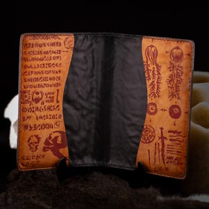 May include: A brown leather wallet with a black spine. The wallet's exterior features a distressed design with red and black symbols and text. The wallet is open, revealing the interior.