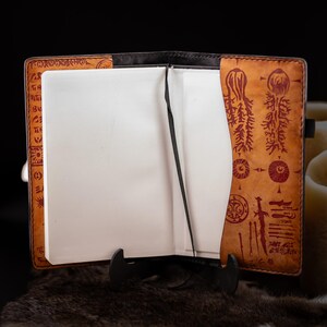 May include: Open leather journal with blank white pages and a black ribbon bookmark. The cover is tan with dark brown edges and red etched designs. The journal is displayed on a black stand.