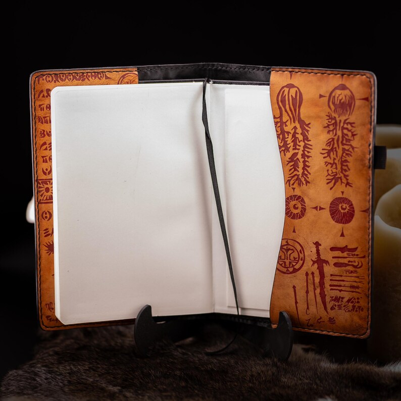 May include: Open leather journal with blank white pages and a black ribbon bookmark. The cover is tan with dark brown edges and red etched designs. The journal is displayed on a black stand.