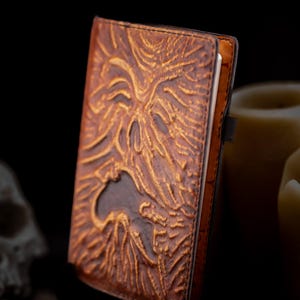 May include: A brown leather journal with an embossed design. The cover features a detailed, textured image of a face with intricate patterns. The journal has a black elastic band and a spine. The overall aesthetic is vintage and gothic.