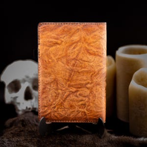 May include: A brown leather journal with a textured surface is displayed on a black stand. The journal is the focal point, with a skull and candles in the blurred background. The leather has a unique, wrinkled pattern.