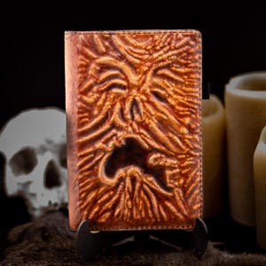 May include: A brown leather-bound book with a carved face design. The cover has a textured, aged appearance with a dark, open mouth. The book is displayed on a black stand, with a skull and candles in the background.