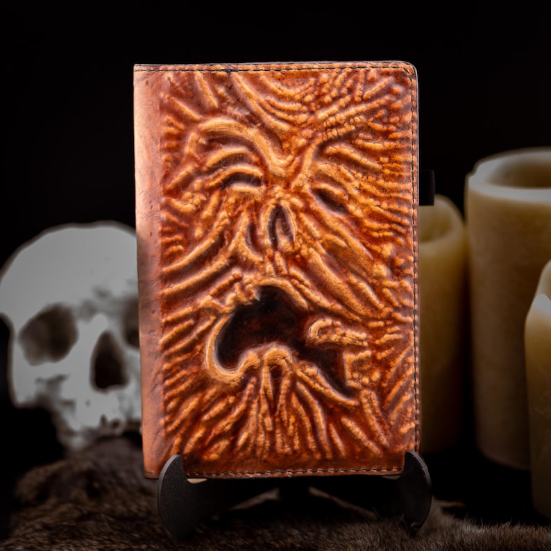 May include: A brown leather-bound book with a carved face design. The cover has a textured, aged appearance with a dark, open mouth. The book is displayed on a black stand, with a skull and candles in the background.